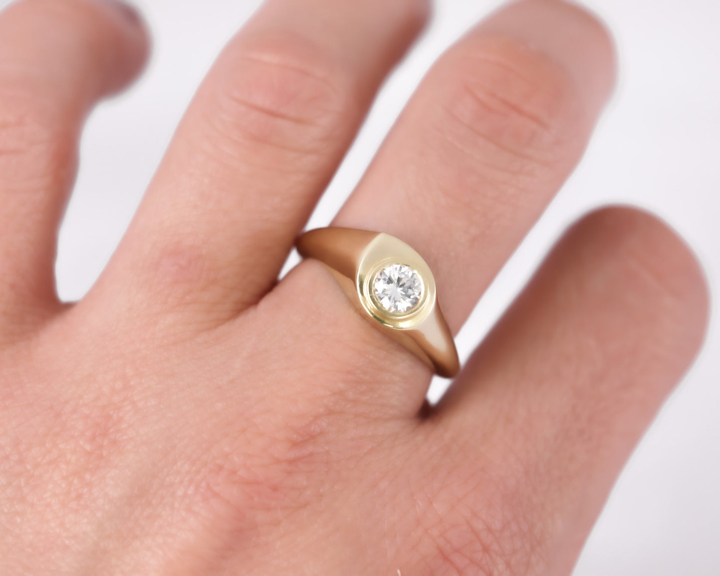 Close-up of a 14K yellow gold bezel-set diamond signet ring worn on a finger with a soft white background