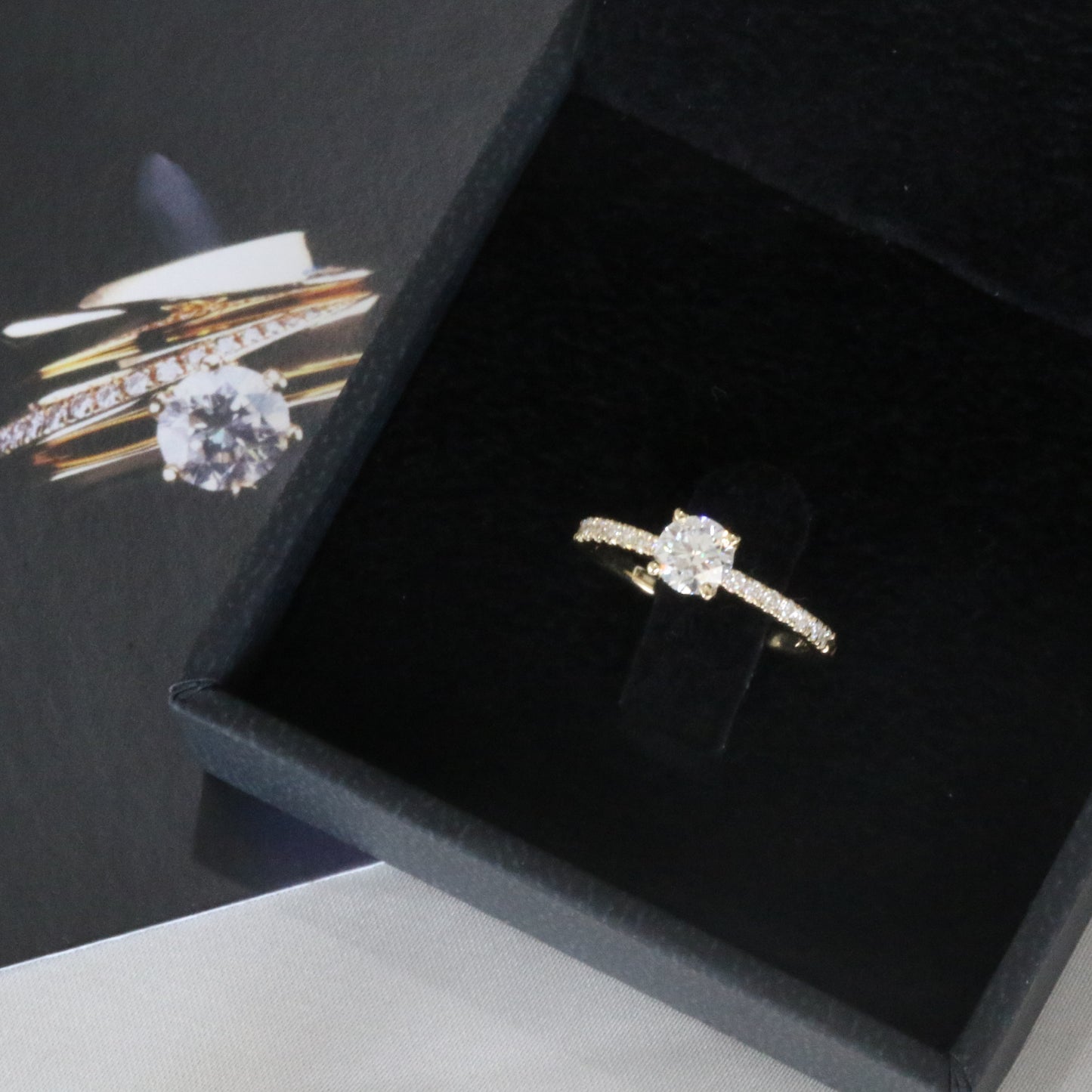 diamond solitaire ring with pave setting on shank inside black jewelry box. card with image of rings under the box