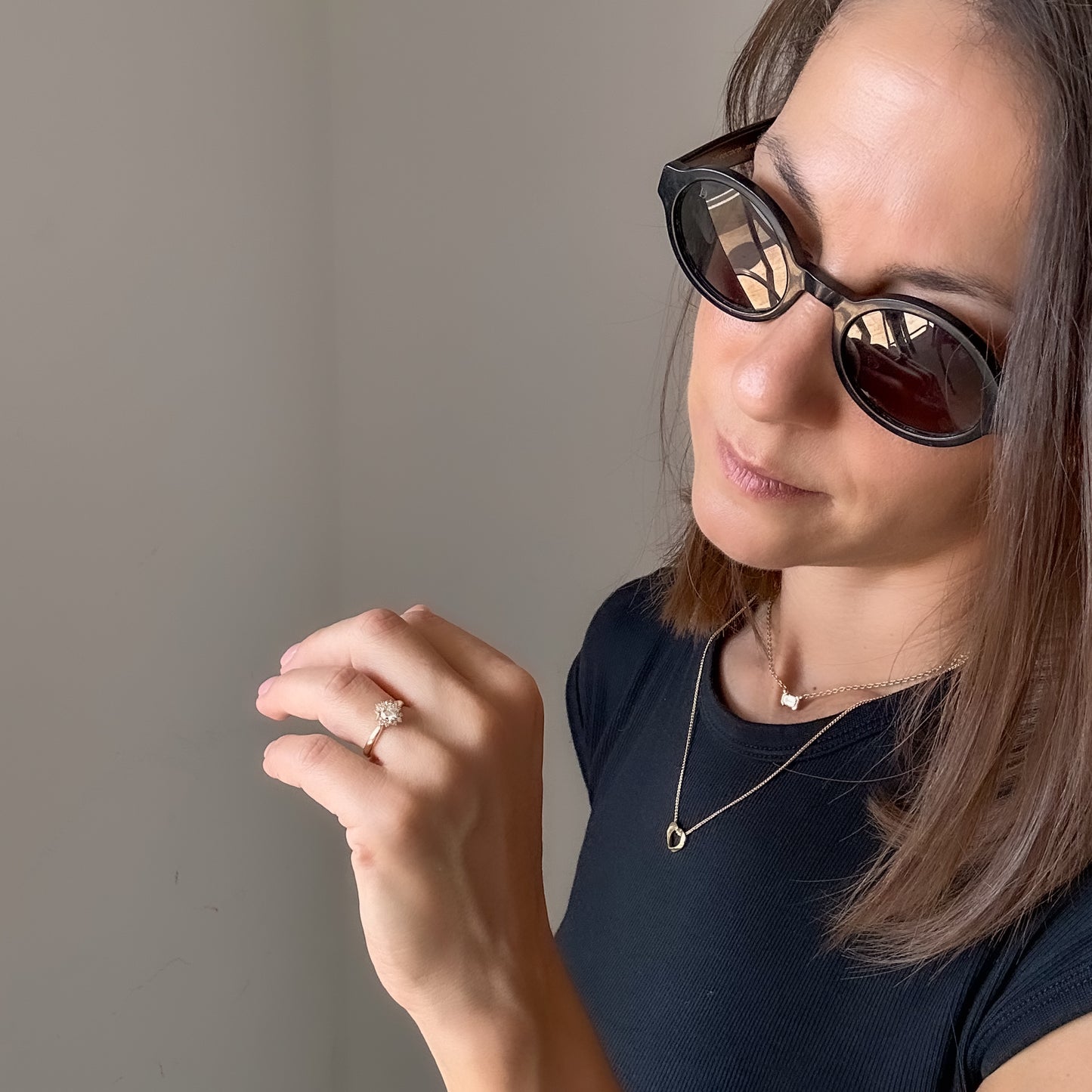 A women with sunglasses looking at her hand wearing a floral engagement ring with a central diamond and smaller surrounding diamonds set in a 14K gold band in a black velvet box.