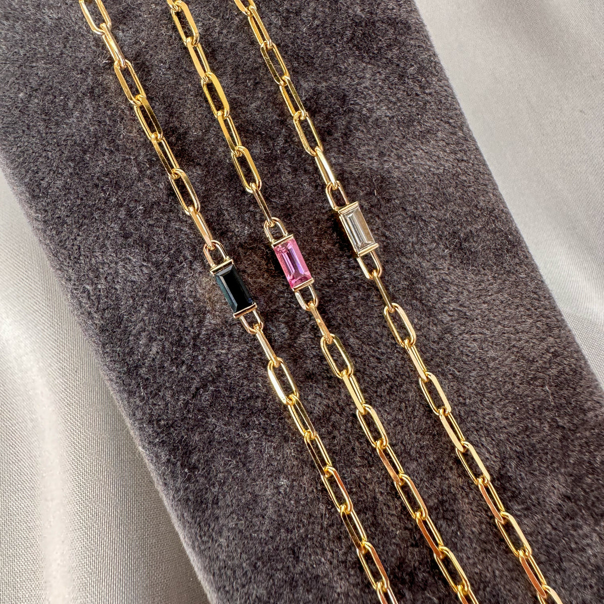 Three gold paperclip chain bracelets with baguette cut pink sapphire, blue sapphire and diamond on a gray velvet stand