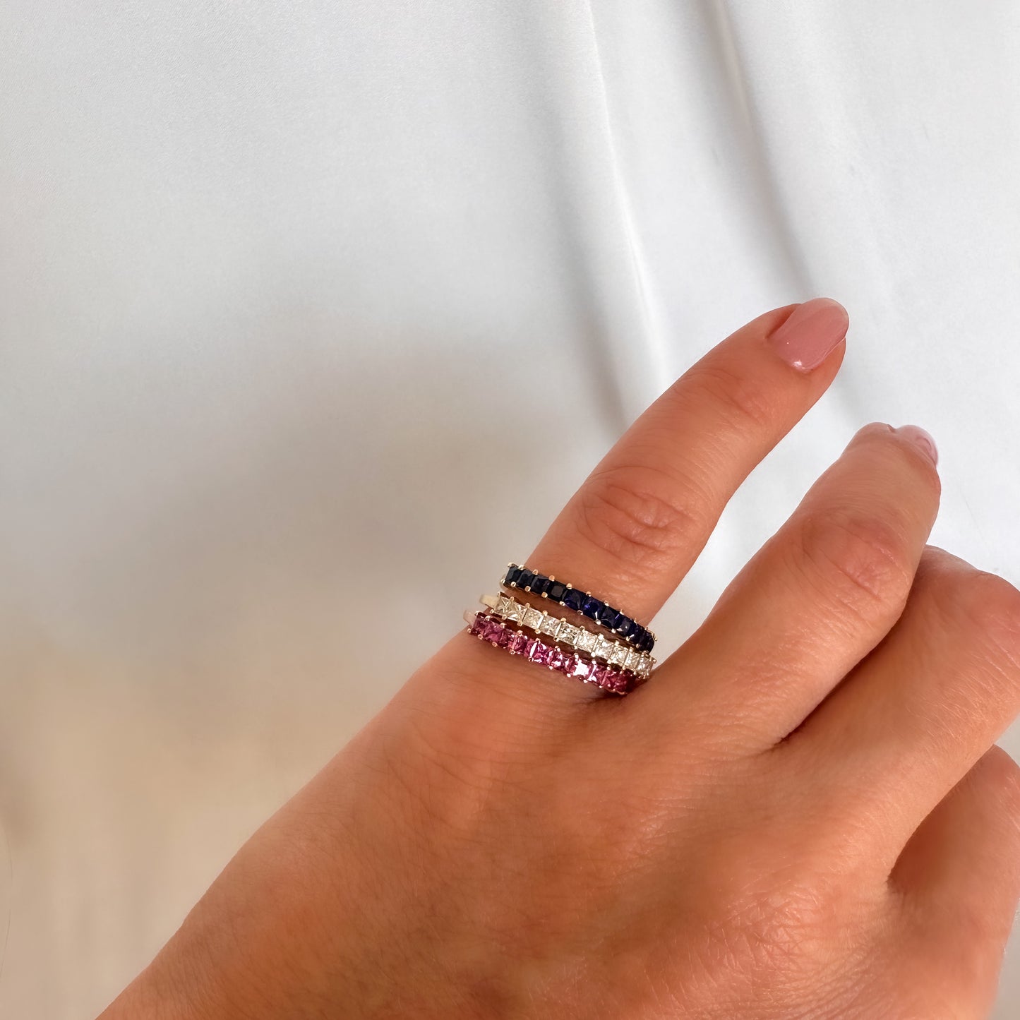 womens hand displaying on pinky finger three princess cut eternity each one with different stones. diamonds, pink sapphires and blue sapphires.
