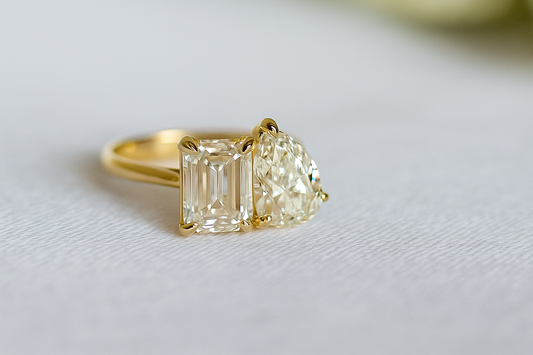 Close-up of a two-stone diamond ring set in 18K yellow gold, featuring emerald and pear-cut diamonds on a soft neutral background. Elegant fine jewelry photography for engagement and diamond education blog.