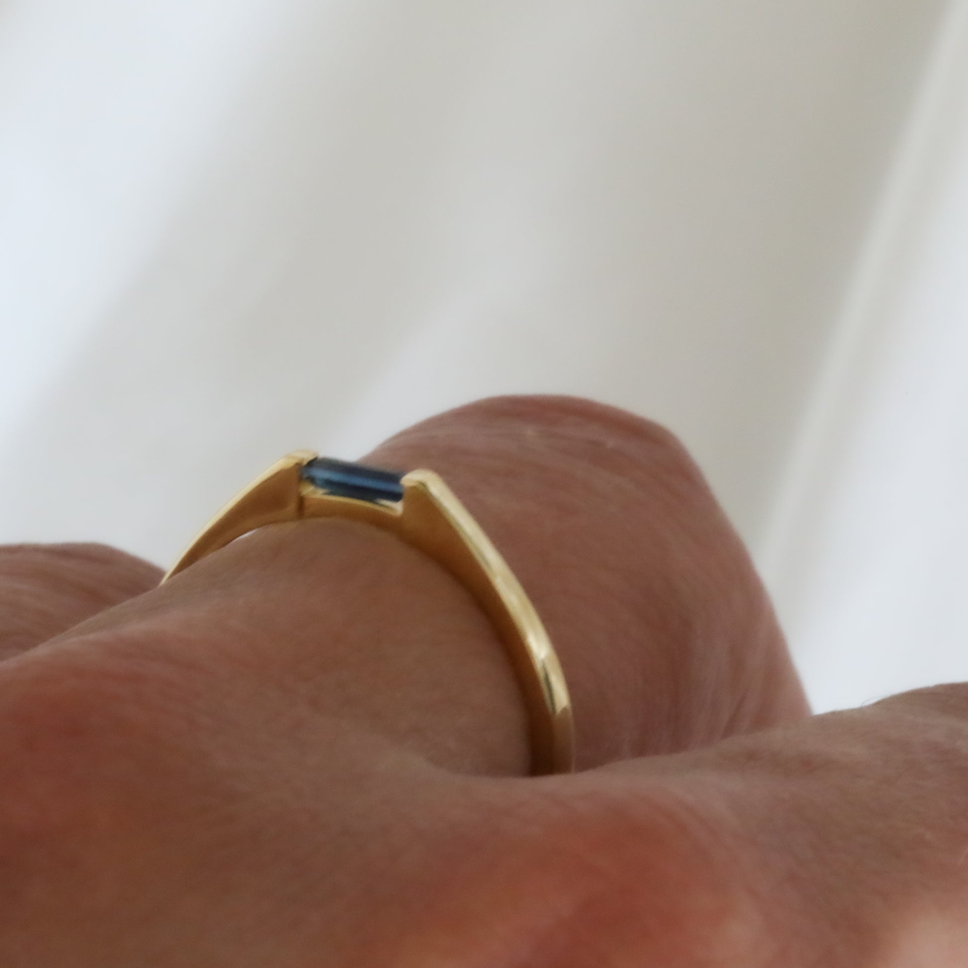 A minimalist style ring with a needle baguette cut blue sapphire set in a 14K yellow gold band, modeled on womens hand against a white background.