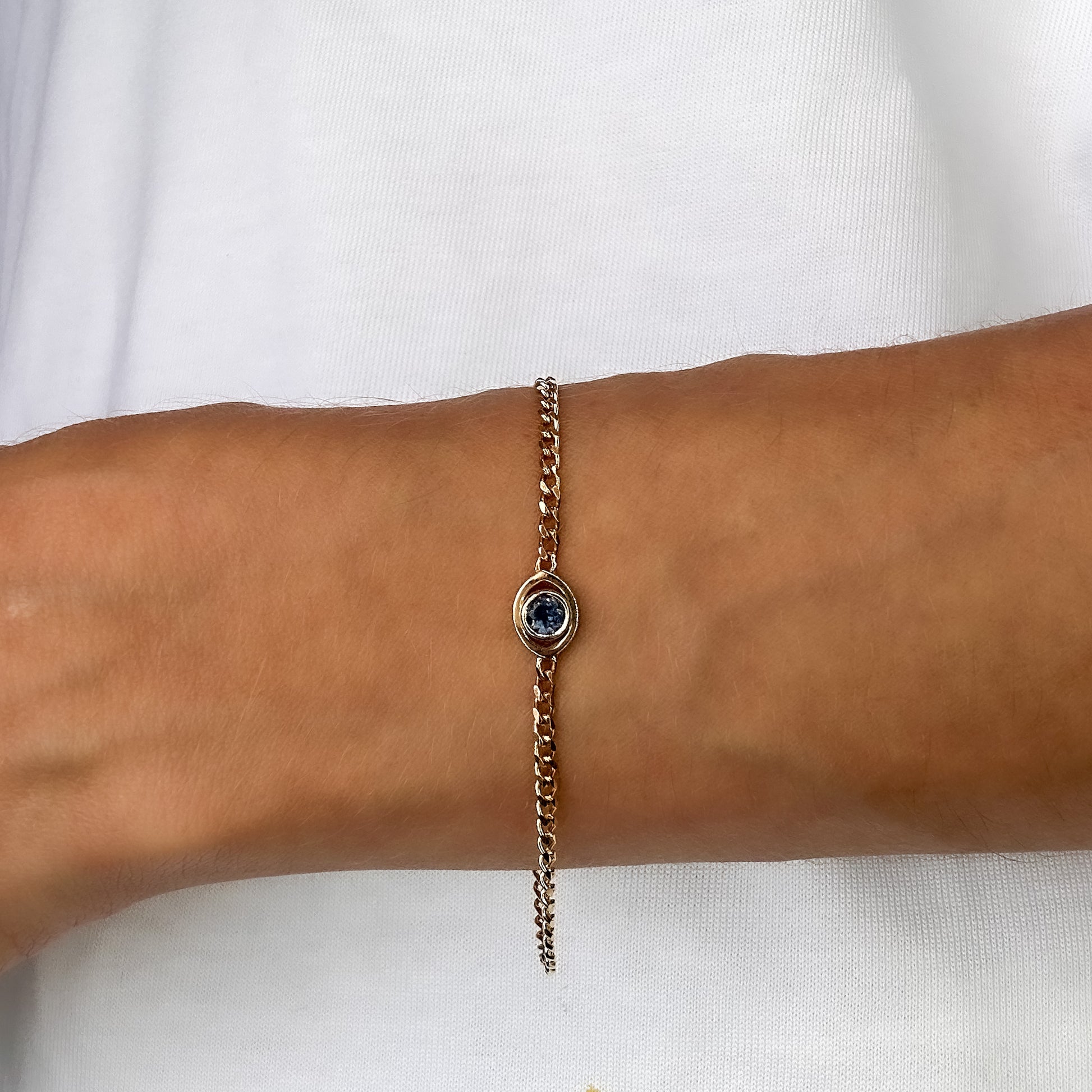 A person wearing a gold curb chain bracelet with a blue sapphire set in a evil eye centerpiece on their wrist.