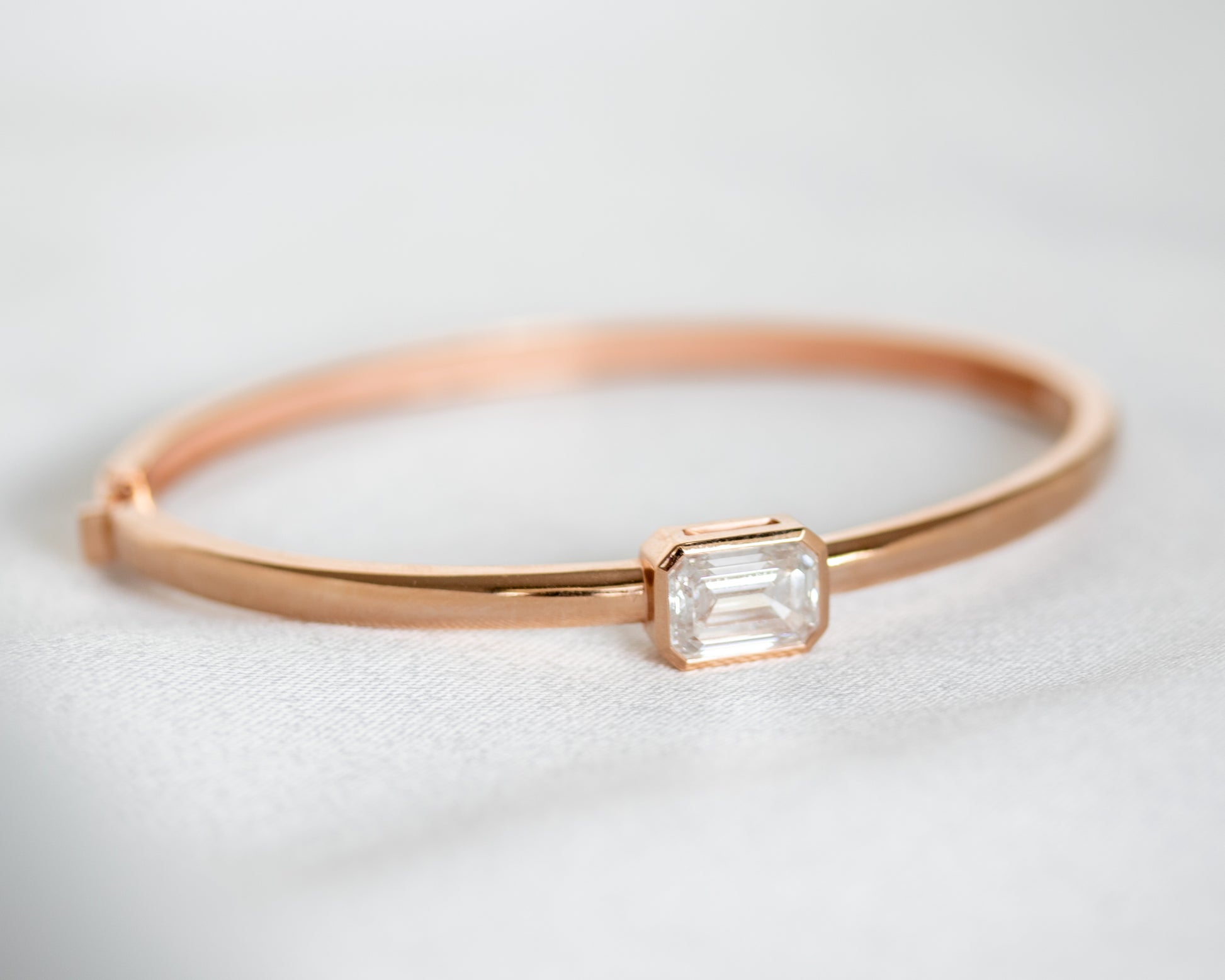 Diagonal view of 14K gold bezel-set emerald cut diamond bangle on a light gray background