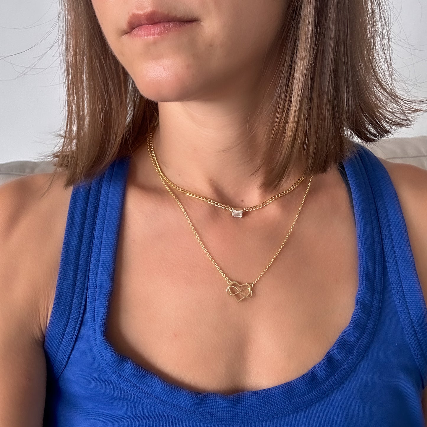 A person wearing a 14K gold diamond solitaire necklace with an emerald cut natural diamond pendant layered with a 14k gold heart pendant necklace.