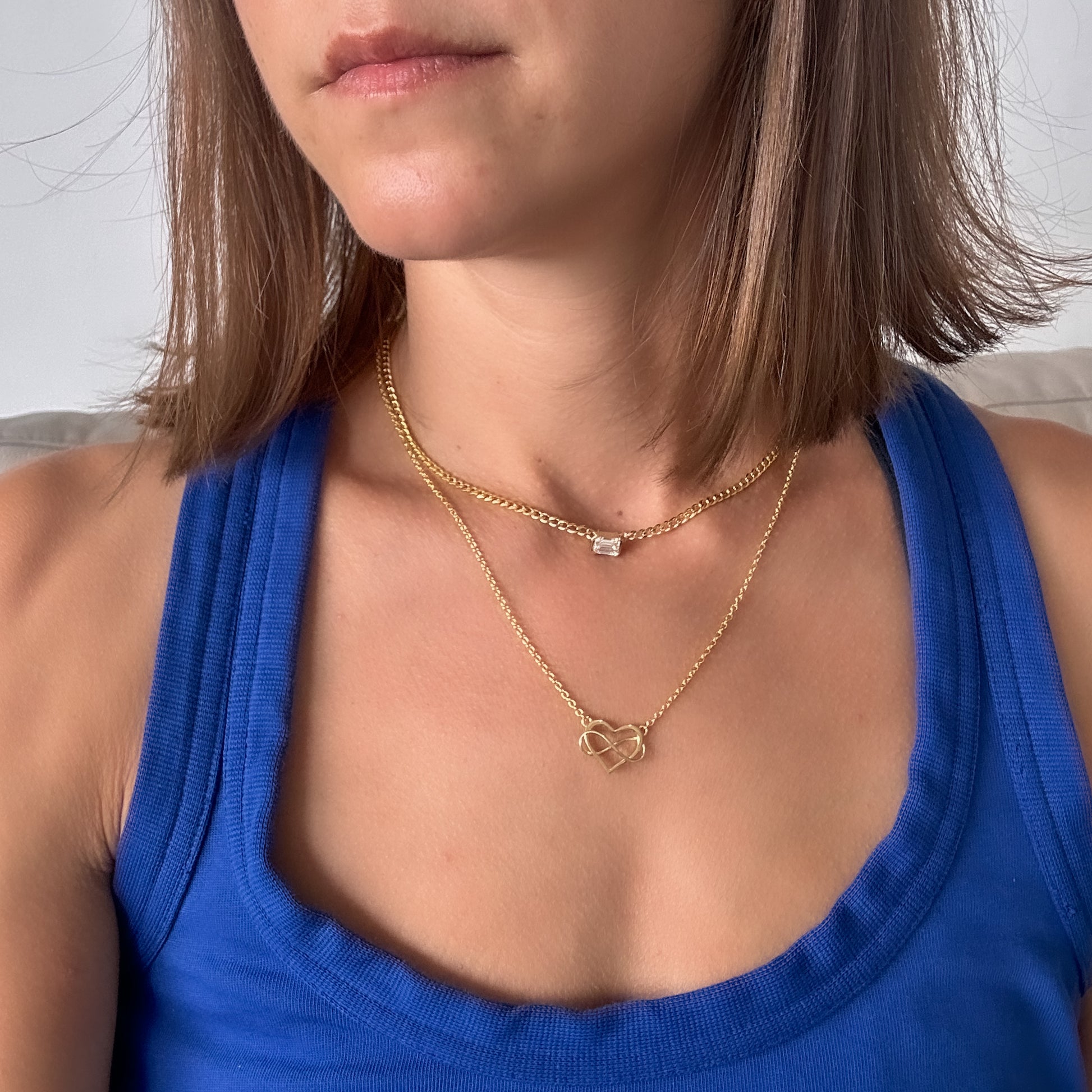 A person wearing a 14K gold diamond solitaire necklace with an emerald cut natural diamond pendant layered with a 14k gold heart pendant necklace.