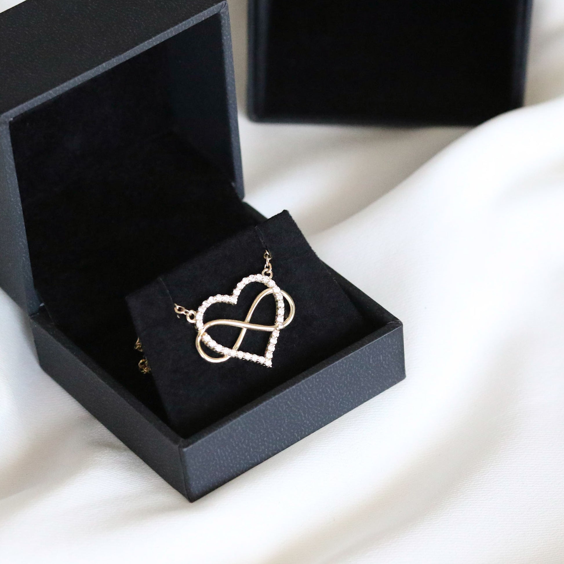 A heart-shaped pendant necklace with a diamonds and gold infinity symbol, attached to a yellow gold chain, displayed black velvet box on white background