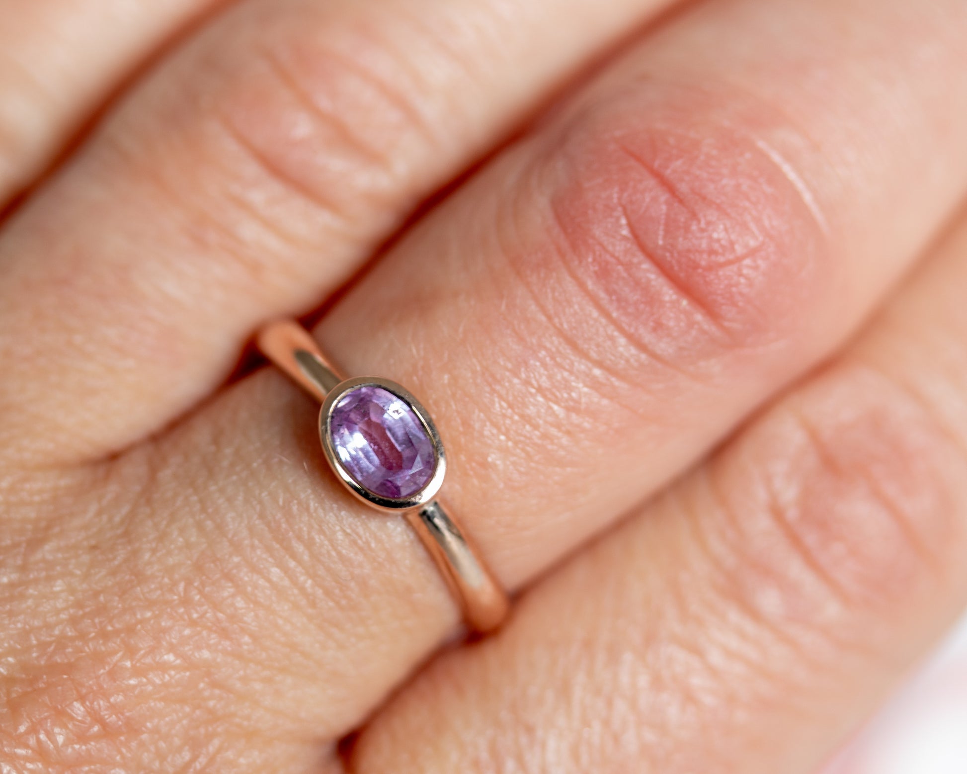 Close-up of bezel-set oval pink sapphire ring on hand, 14K gold