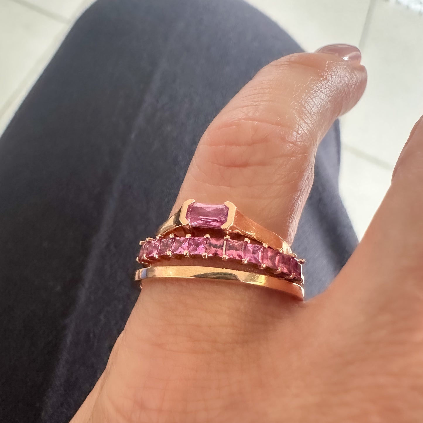 womens pinky finger displaying three rose gold rings. one with emerald cut pink sapphire, one eternity ring with pink princess cut sapphires and one solid rose gold minimalist band.