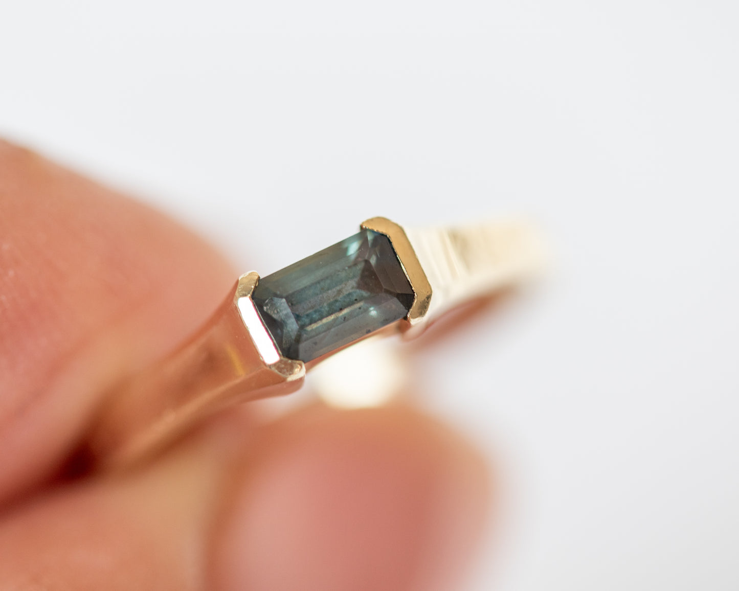 Gold ring with emerald cut sapphire held by a hand against a white background