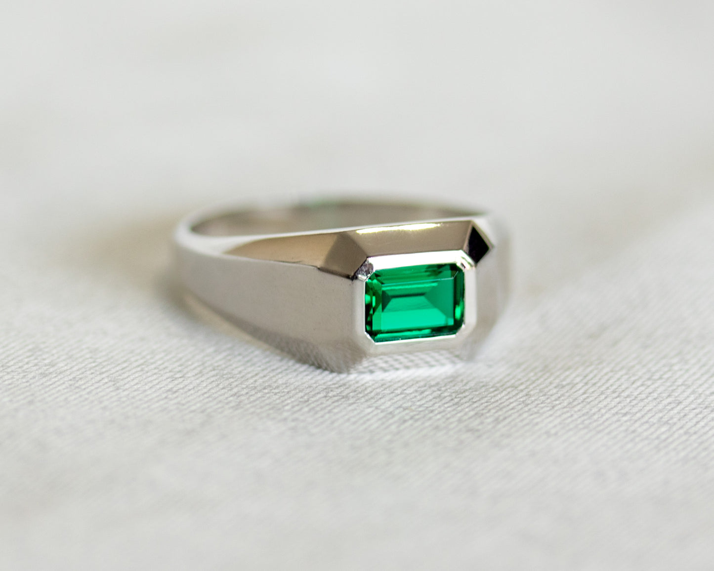 White gold geometric signet ring with emerald-cut Chatham emerald on soft fabric background