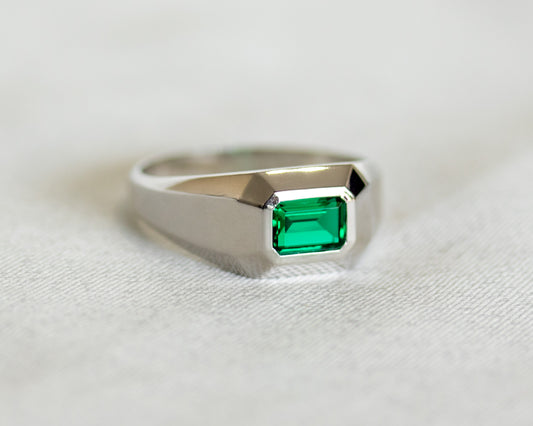 White gold geometric signet ring with emerald-cut Chatham emerald on soft fabric background