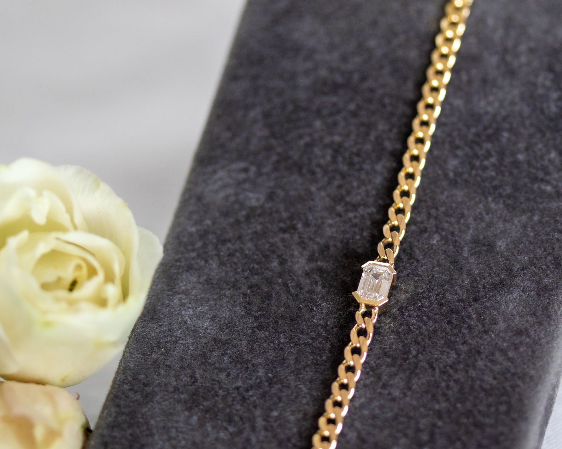 Gold chain bracelet with an emerald cut diamond on a gray surface next to a white rose.