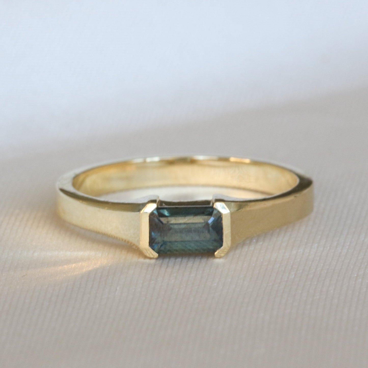 A yellow gold ring with a emerald cut natural sapphire stone, set in a half bezel setting on gray background