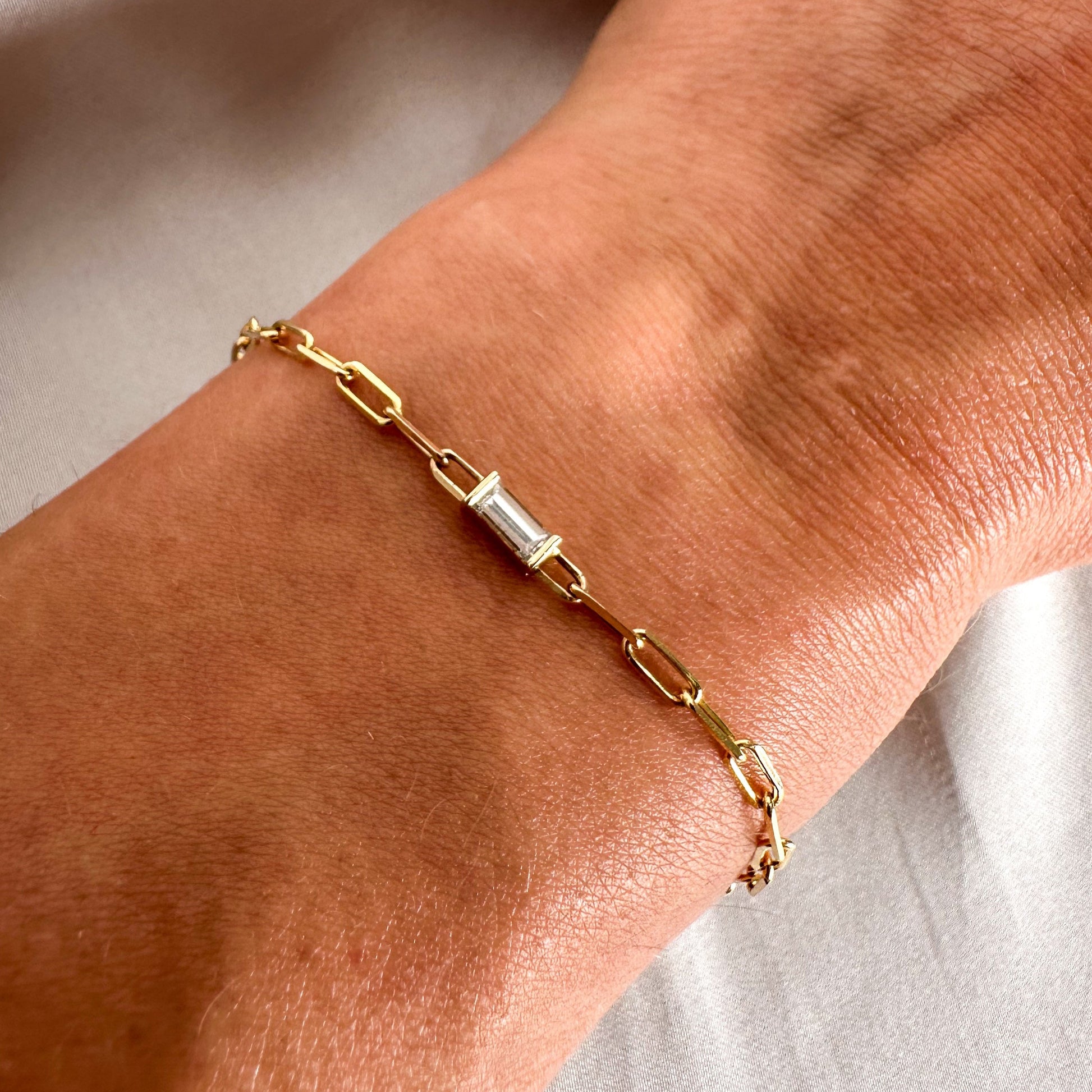 A person's wrist wearing a gold bracelet with a baguette cut paper clip chain and a small baguette cut diamond.