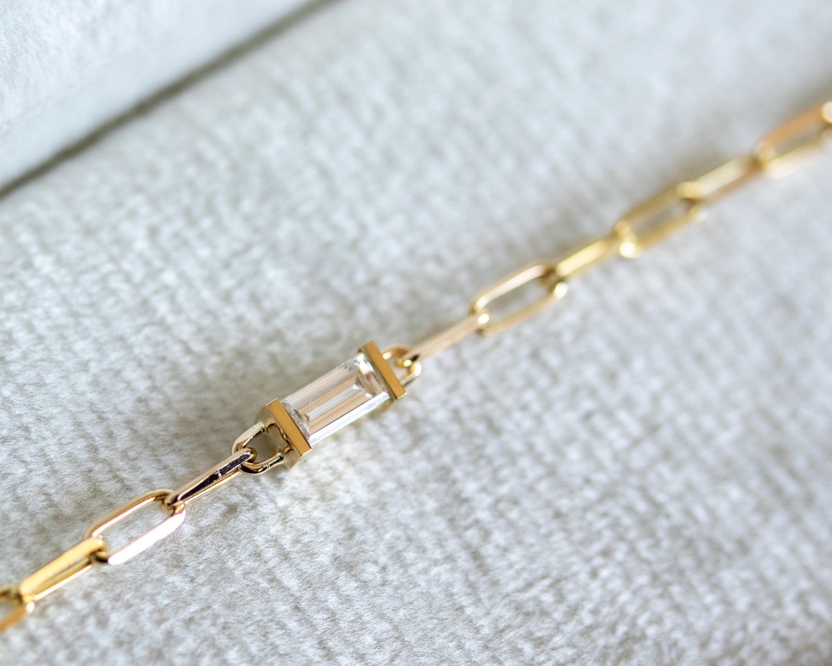 Baguette cut diamond in a half bezel on a gold paperclip chain, close-up on a textured white surface