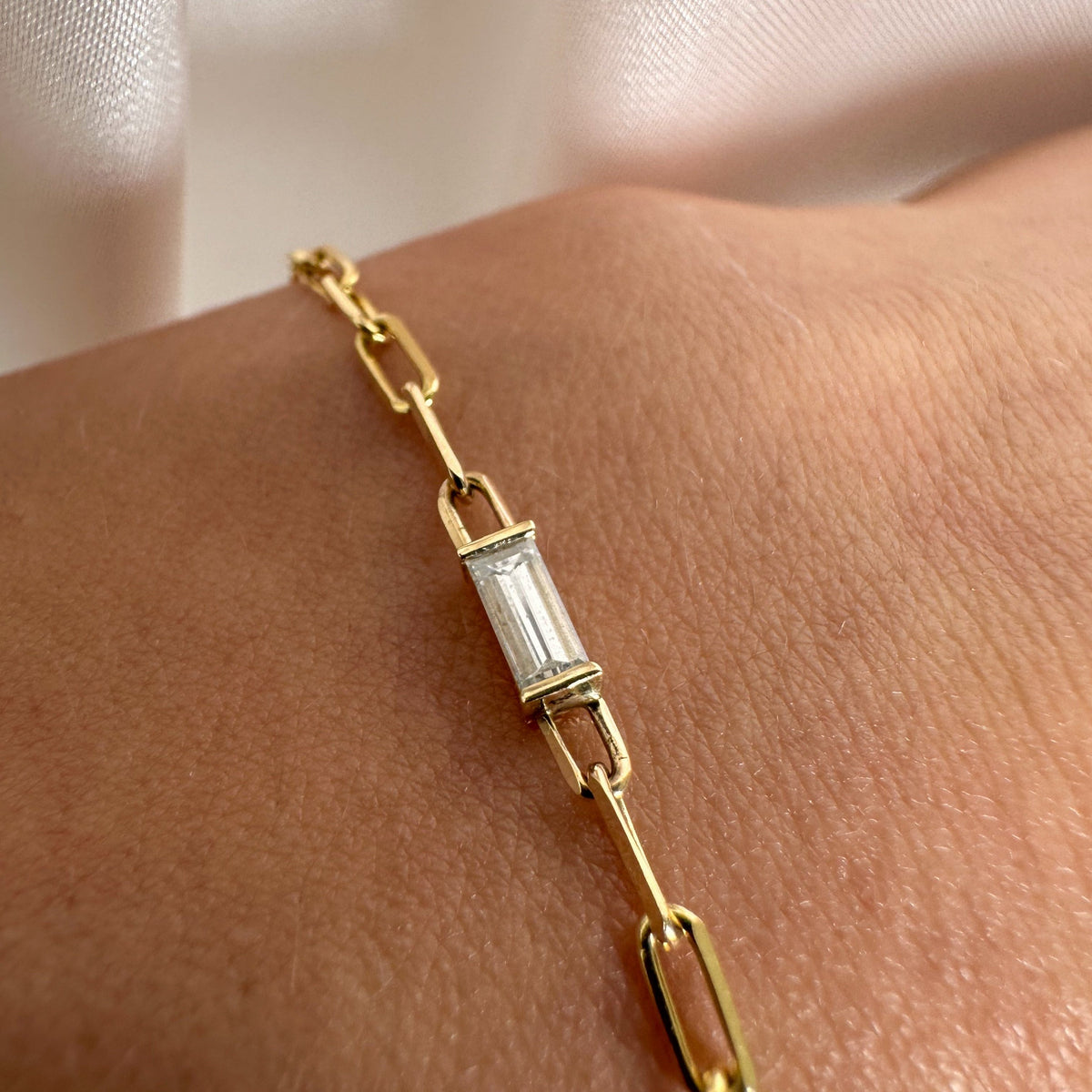 Close-up of a baguette cut diamond in a half bezel on a gold paperclip chain bracelet, worn on the wrist