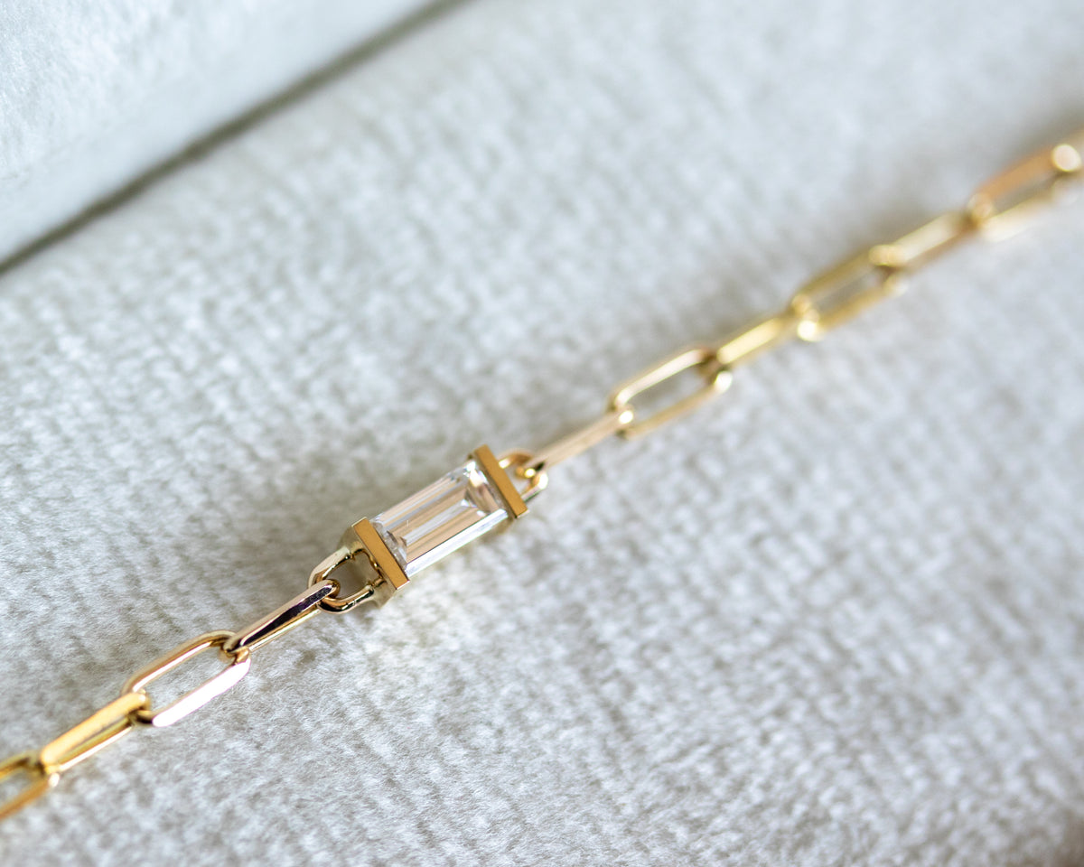 Gold paperclip chain bracelet with a baguette cut diamond on a textured white surface