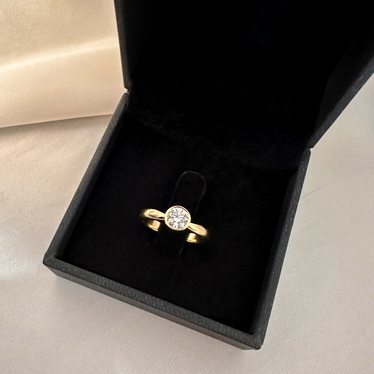 Gold bezel solitaire engagement ring with a round diamond in a black velvet box, full view