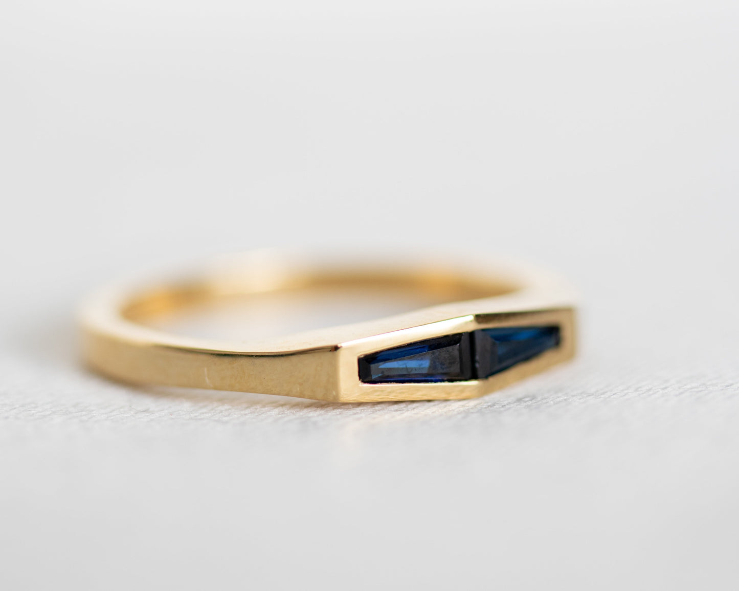 Two tapered baguette blue sapphires bezel set in a 14K yellow gold band, angled close-up on a light grey surface.