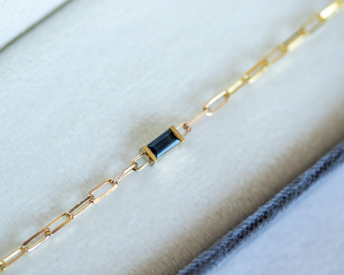 Gold paperclip chain bracelet with a baguette cut blue sapphire on a white background
