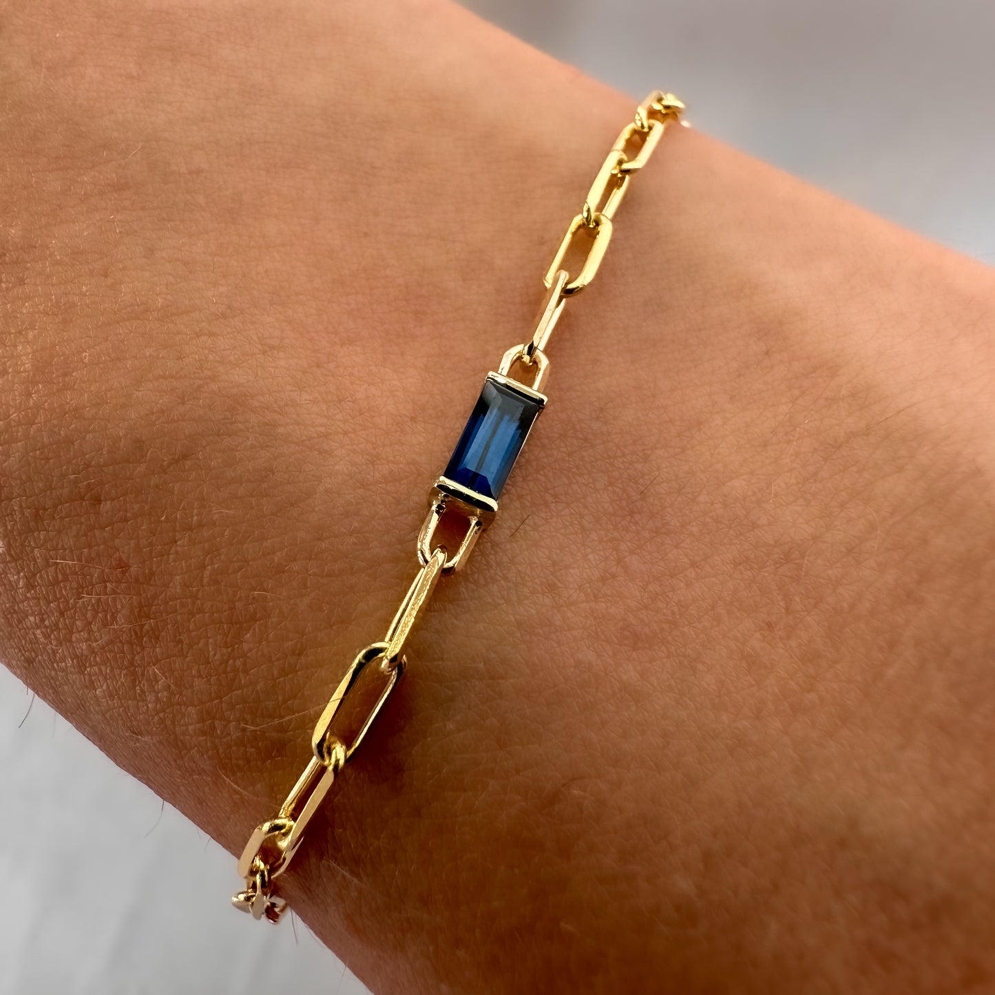 A gold bracelet featuring a baguette cut blue sapphire stone, with a chain of paper clips attached to it, worn on a wrist.