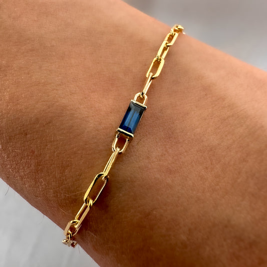 A gold bracelet featuring a baguette cut blue sapphire stone, with a chain of paper clips attached to it, worn on a wrist.