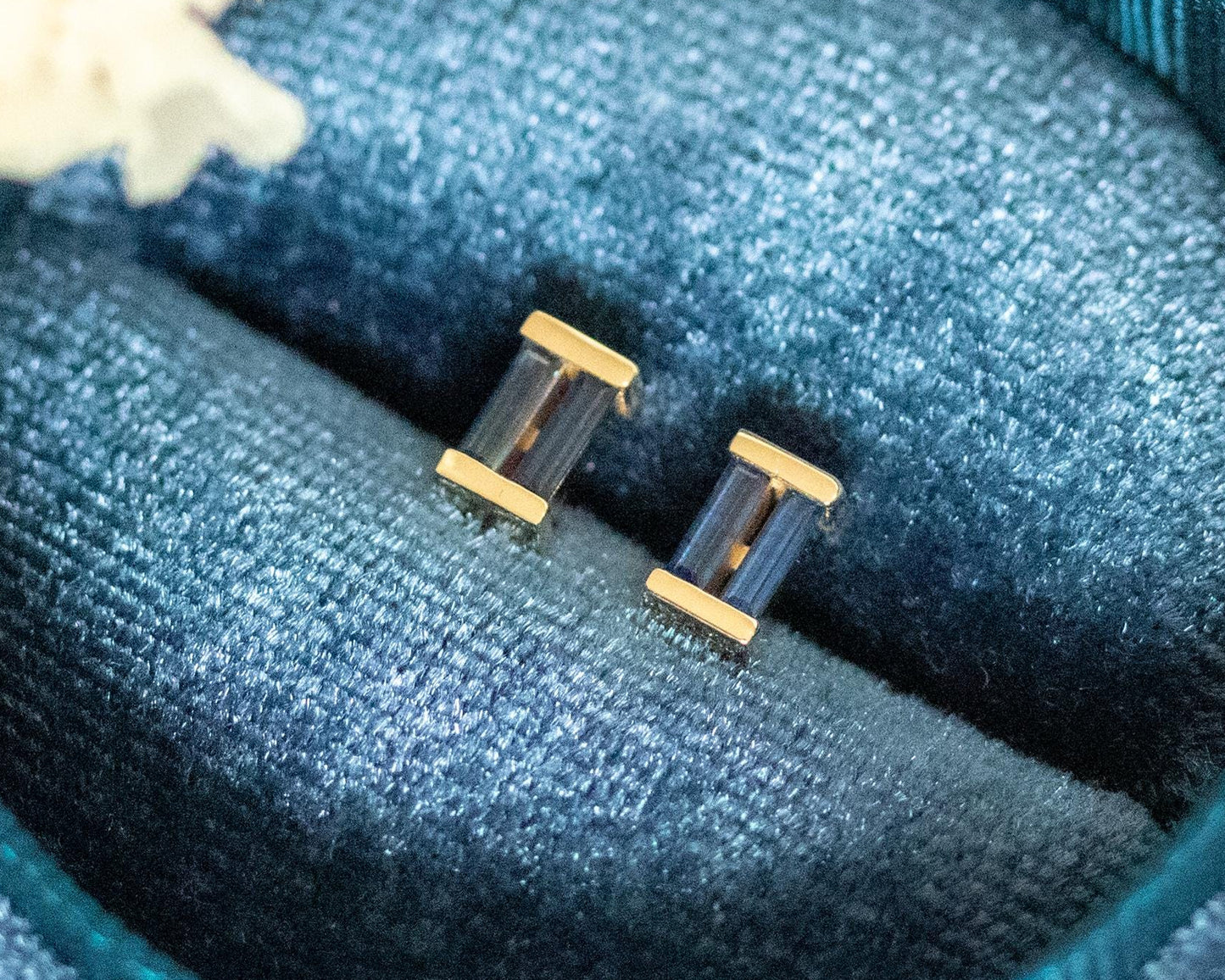 Blue sapphire baguette stud earrings in gold, displayed in a blue velvet box with dried flowers