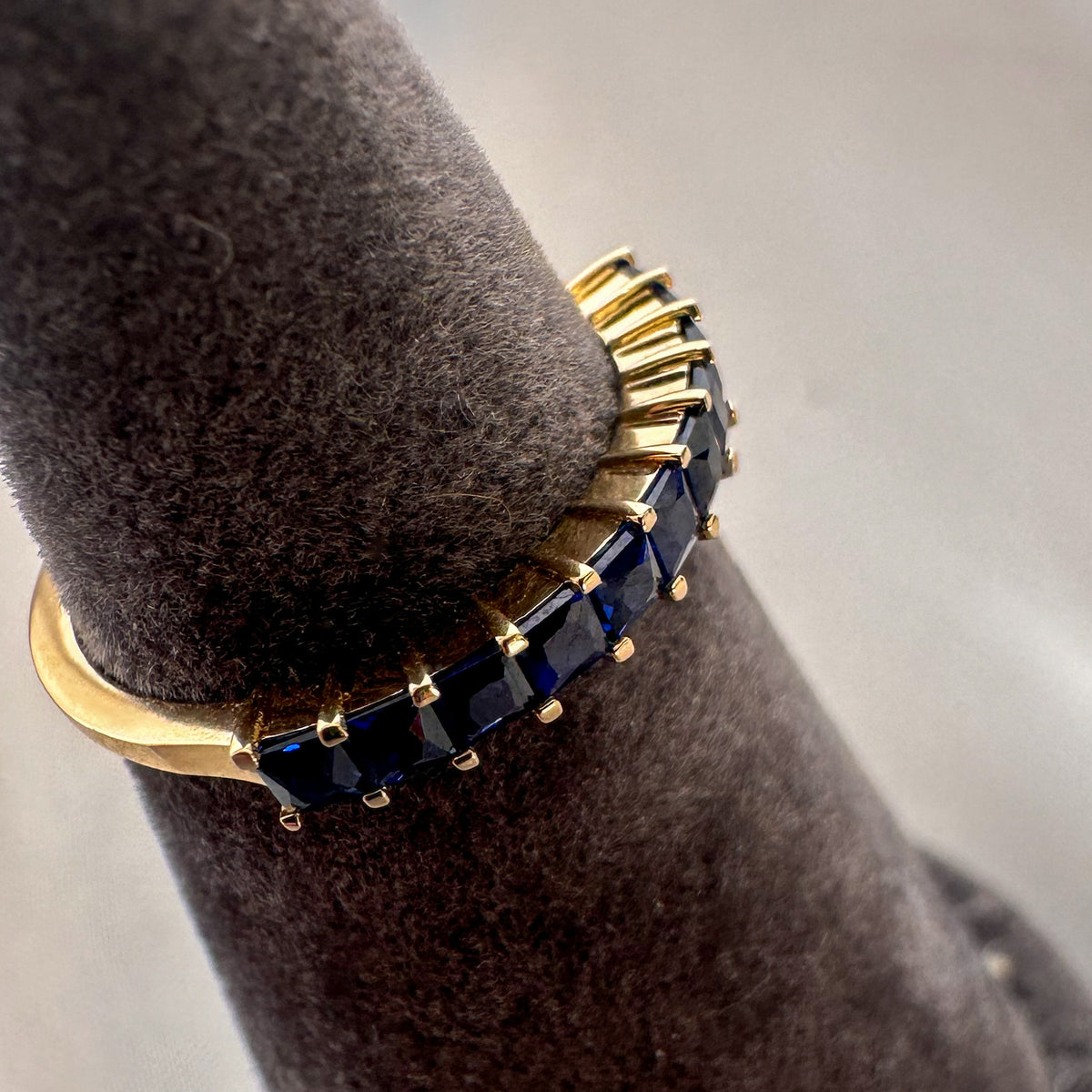 Close-up of princess cut royal blue sapphires in a 14K yellow gold band on a gray ring stand