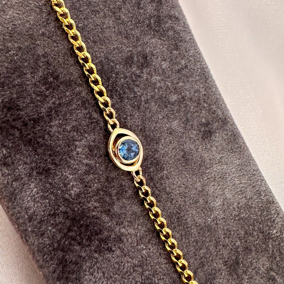 Close-up of a bezel-set blue sapphire evil eye on a 14K yellow gold curb chain, on a gray velvet stand