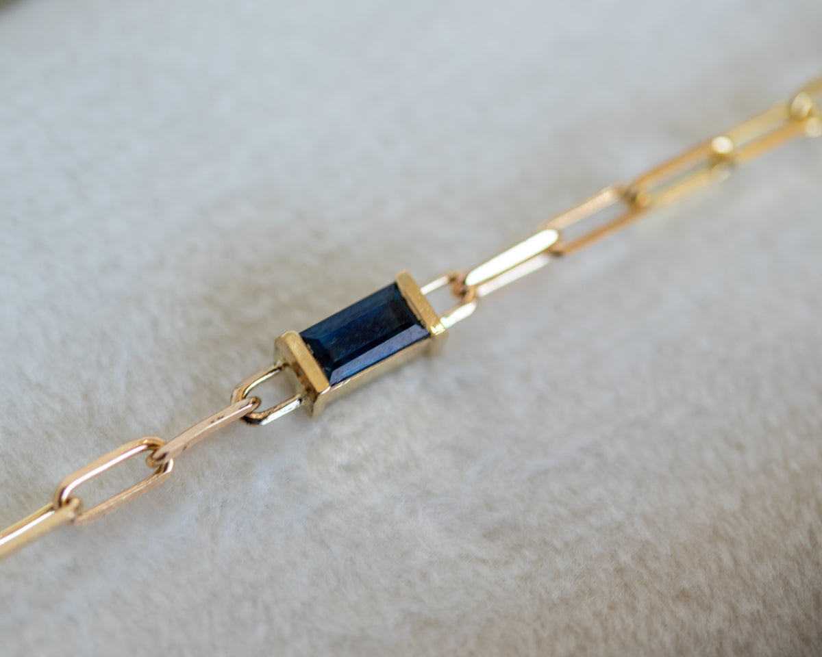 Baguette cut blue sapphire in a half bezel on a gold paperclip chain, on a light gray background