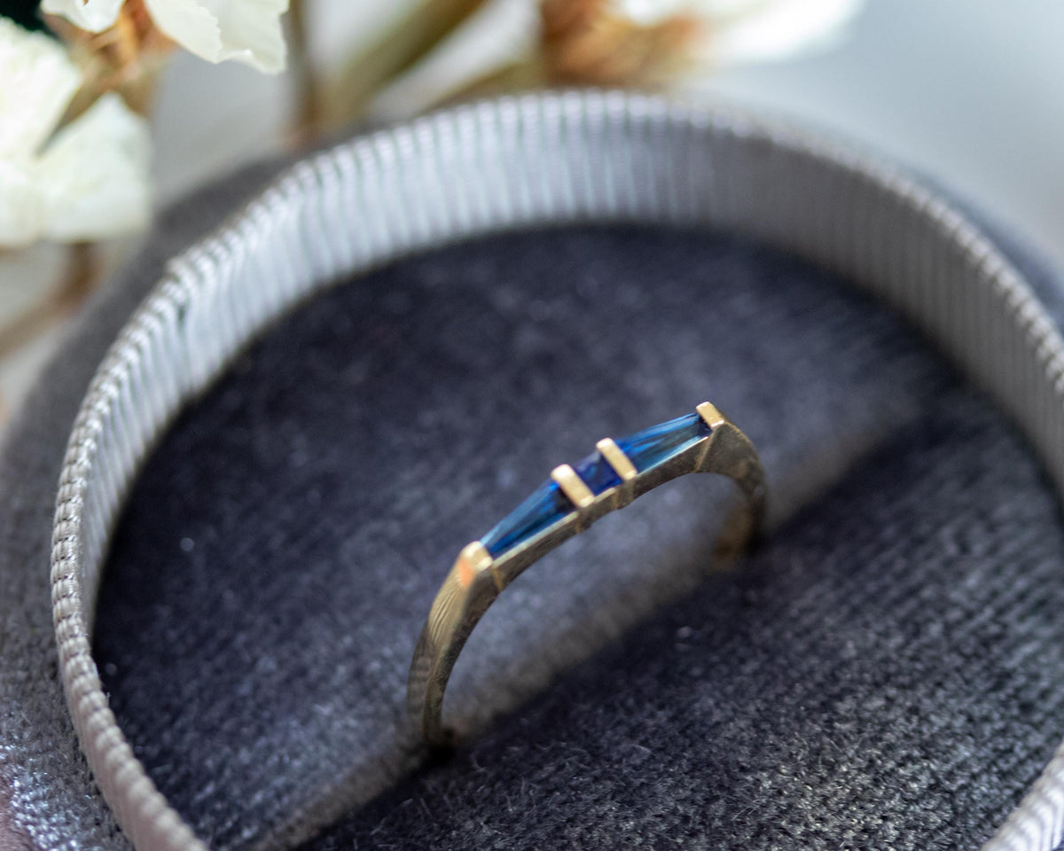 Princess-cut and tapered baguette blue sapphire ring in yellow gold, in a gray velvet ring box with dried flowers