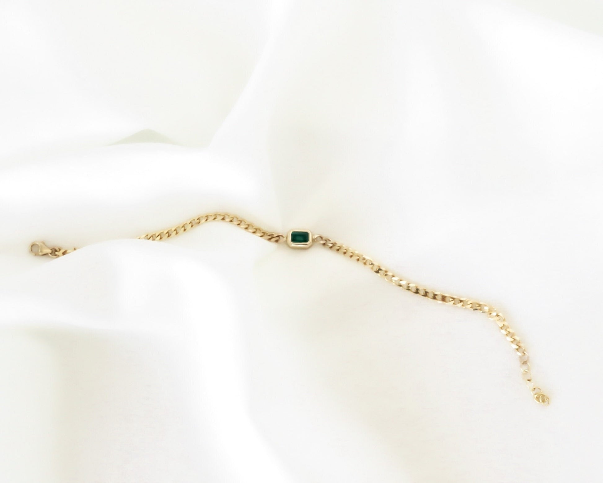 14K yellow gold curb chain bracelet with emerald cut green emerald laid flat on a soft white background