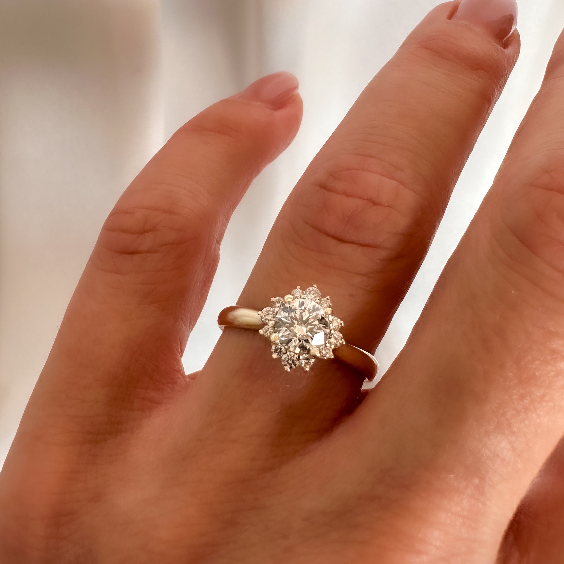 A person's hand displaying a floral engagement ring with a central diamond and smaller surrounding diamonds set in a 14K gold band.
