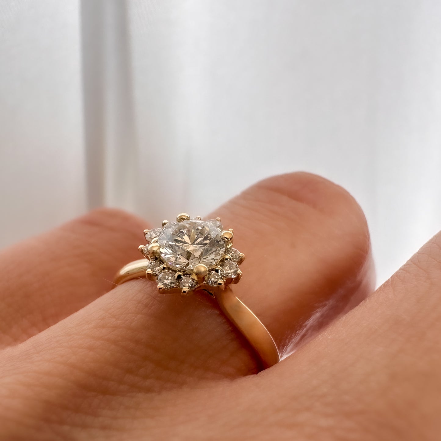close up of a person's hand displaying a floral engagement ring with a central diamond and smaller surrounding diamonds set in a 14K gold band.