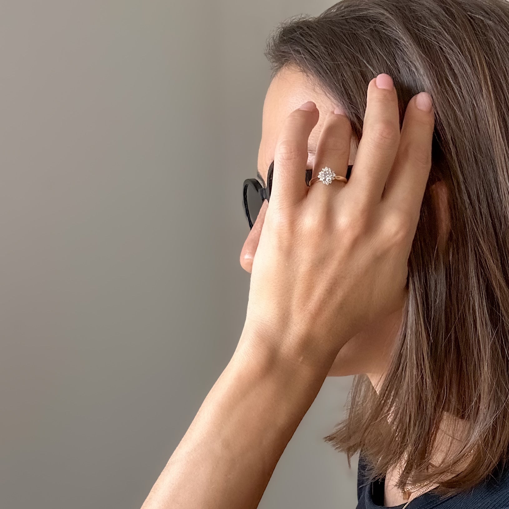 A women with sunglasses stroking her hair and on her hand she is wearing a floral engagement ring with a central diamond and smaller surrounding diamonds set in a 14K gold band in a black velvet box.