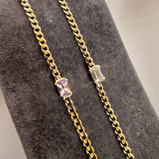 Two 14K gold curb chain bracelets, one with an emerald cut diamond and one with a pink sapphire, on a gray stand