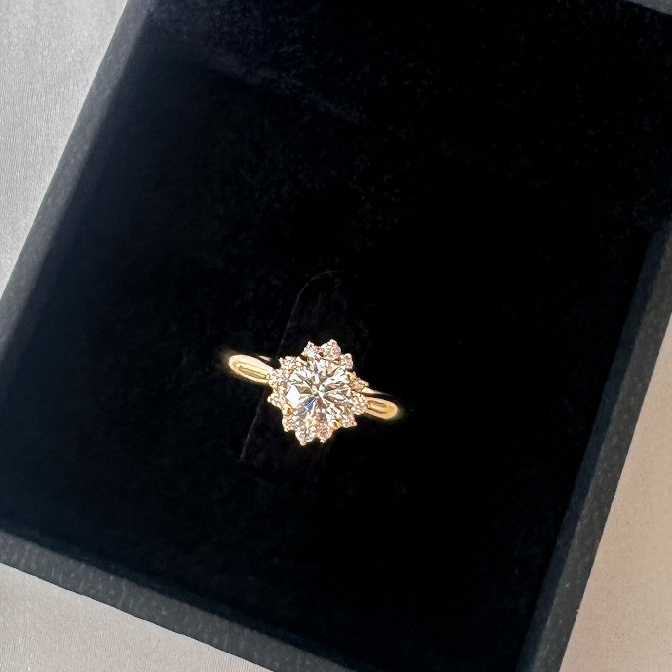 A close up of a floral engagement ring with a central diamond and smaller surrounding diamonds set in a 14K gold band in a black velvet box.