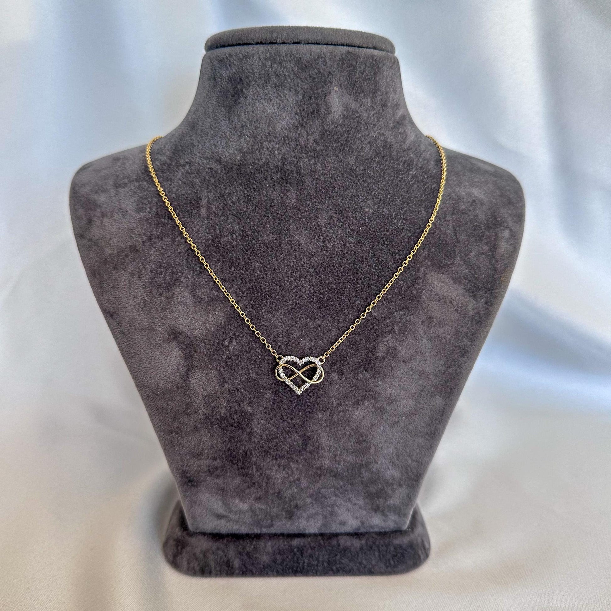 Gold necklace with a heart and infinity symbol on a gray velvet stand.