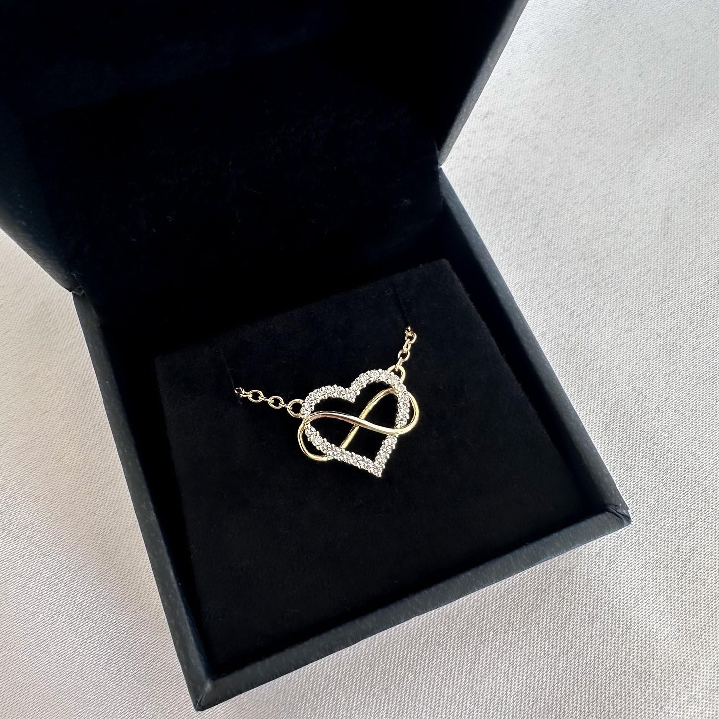 Gold necklace with heart and infinity symbol in a black jewelry box on a light gray surface