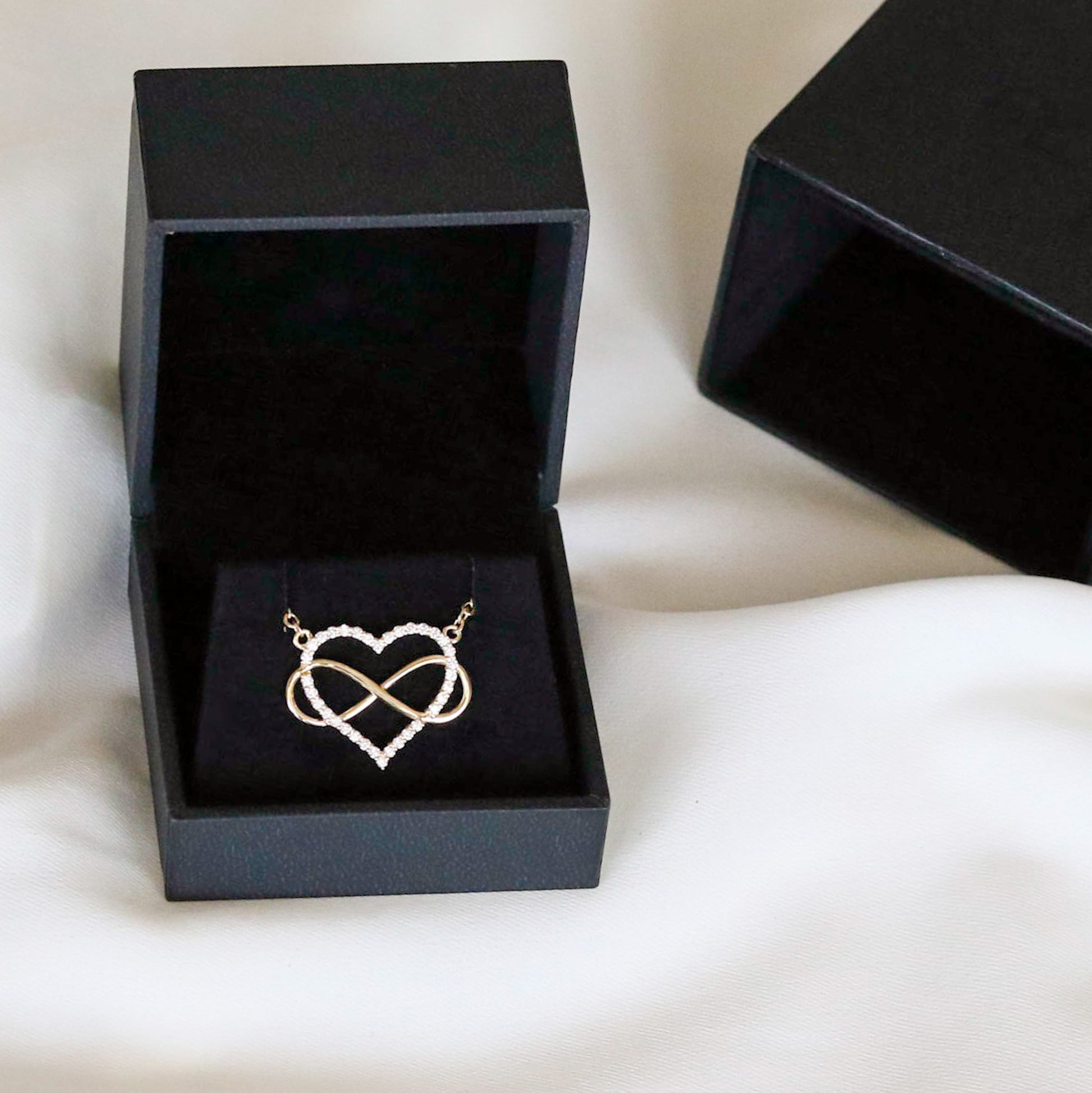 A heart-shaped pendant necklace with a diamonds and gold infinity symbol, attached to a yellow gold chain, displayed black velvet box on white background