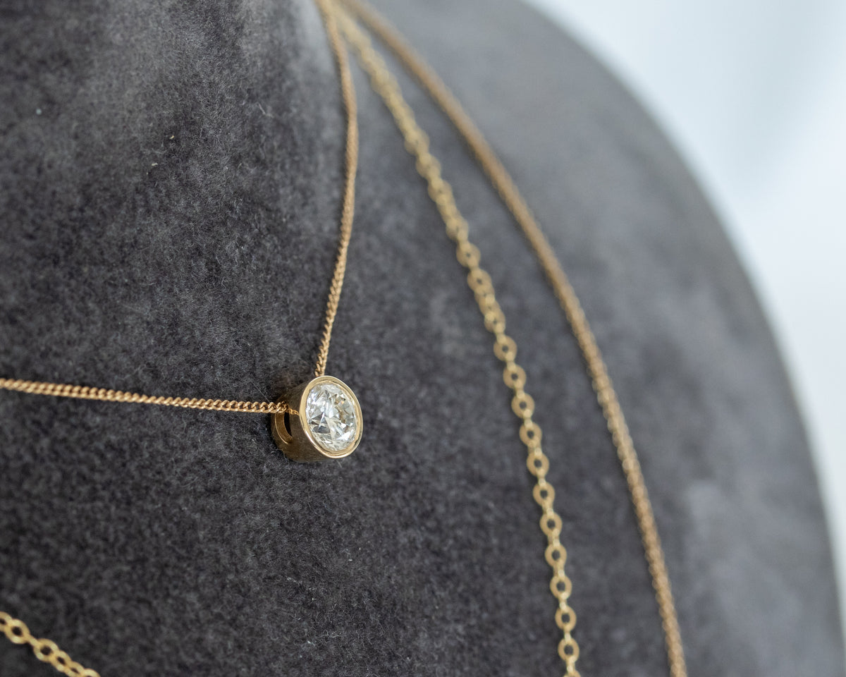 Round diamond bezel pendant on a fine 14K yellow gold cable chain, displayed on a grey jewelry bust.