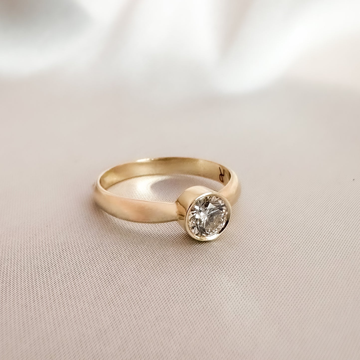 gold solitaire ring with bezel setting on off white background off side view