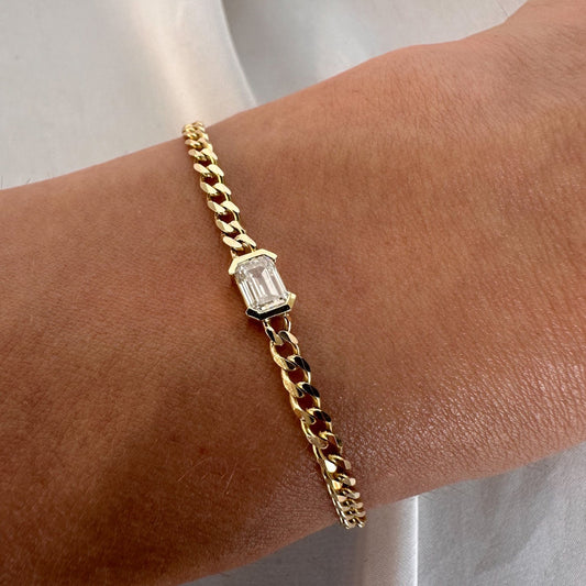 A gold bracelet with a curb chain design featuring an emerald cut diamond solitaire, displayed on a person's wrist.
