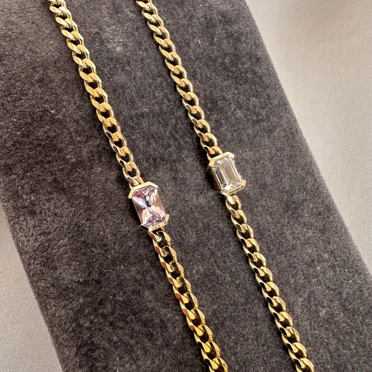 Two gold bracelets with a curb chain design one featuring an emerald cut diamond solitaire, and the other  featuring an emerald cut natural pink sapphire displayed on a gray textured stand