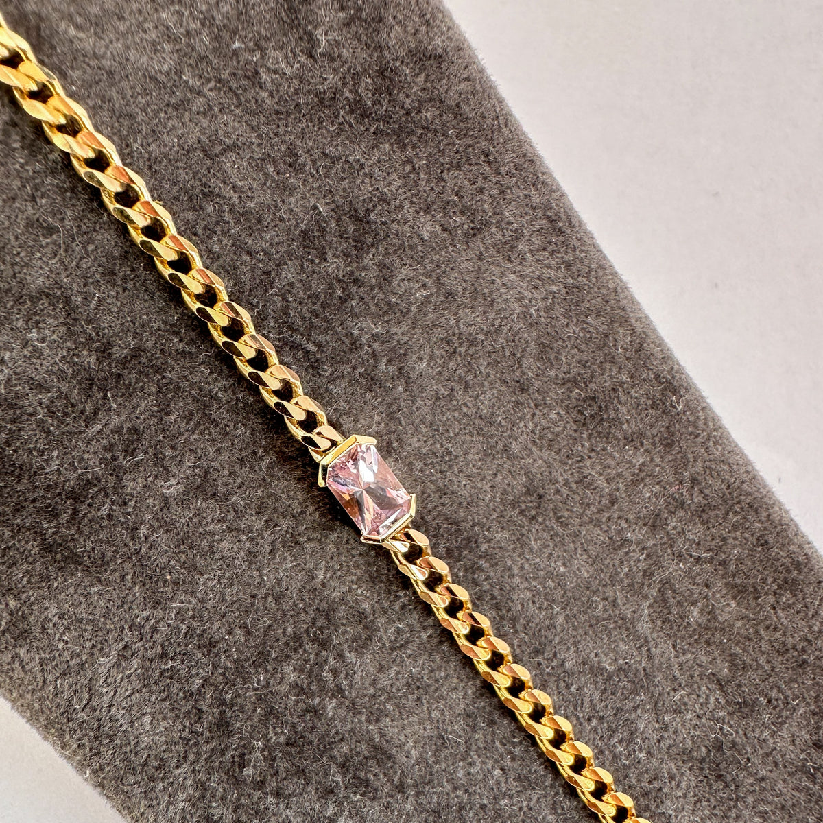 Emerald cut pink sapphire in a half bezel setting on a fine 14K yellow gold curb chain bracelet, close-up on gray velvet