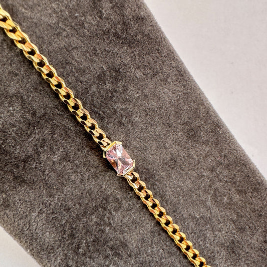Emerald cut pink sapphire in a half bezel setting on a fine 14K yellow gold curb chain bracelet, close-up on gray velvet