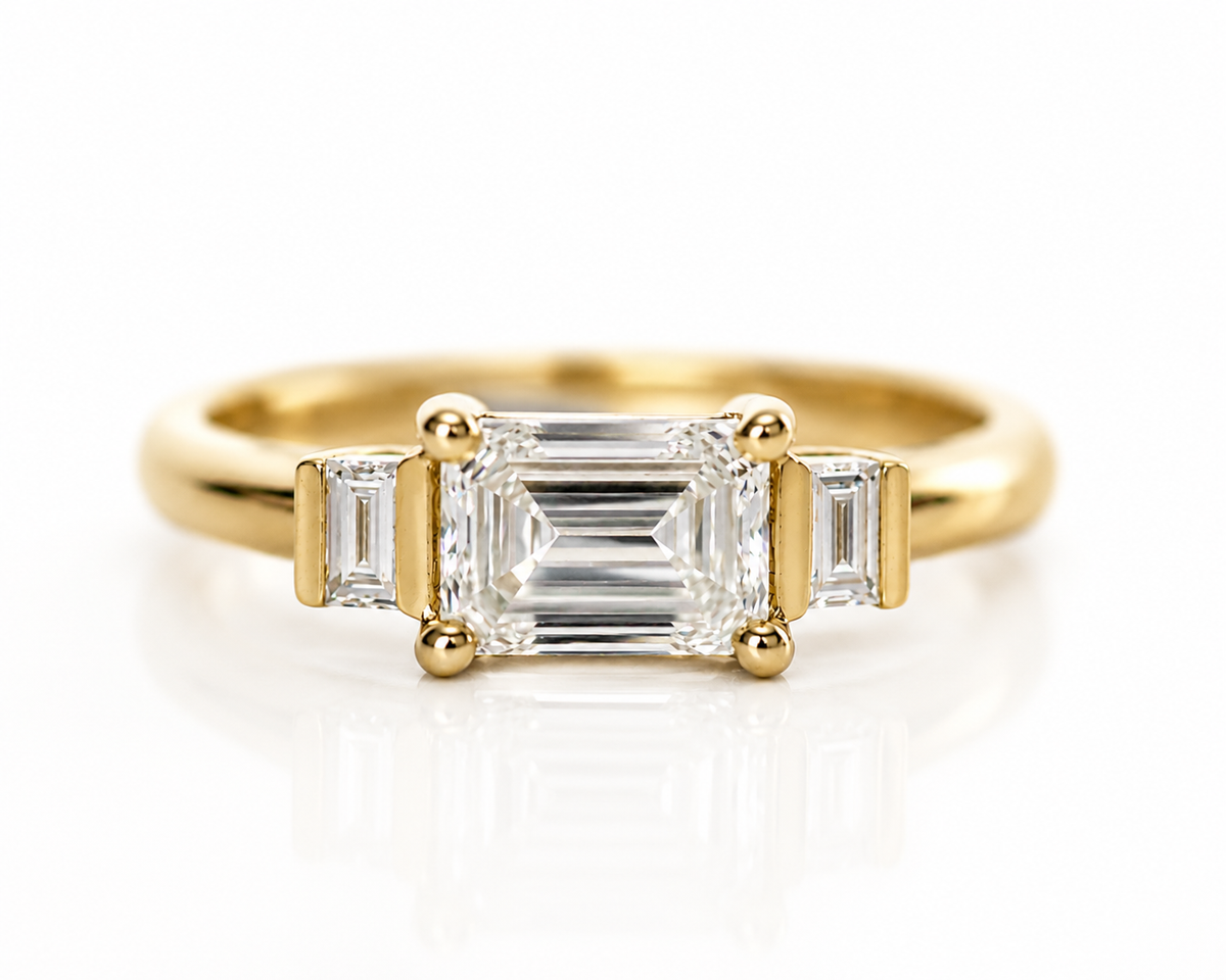 Front view of an emerald cut three-stone engagement ring with baguette side diamonds in yellow gold.