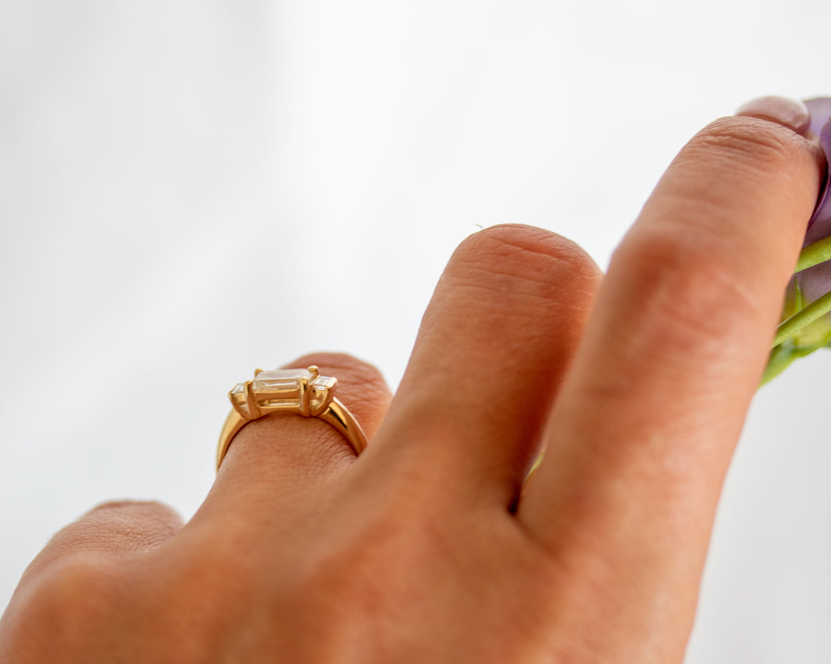 Close-up of an emerald cut three-stone engagement ring worn on the hand.